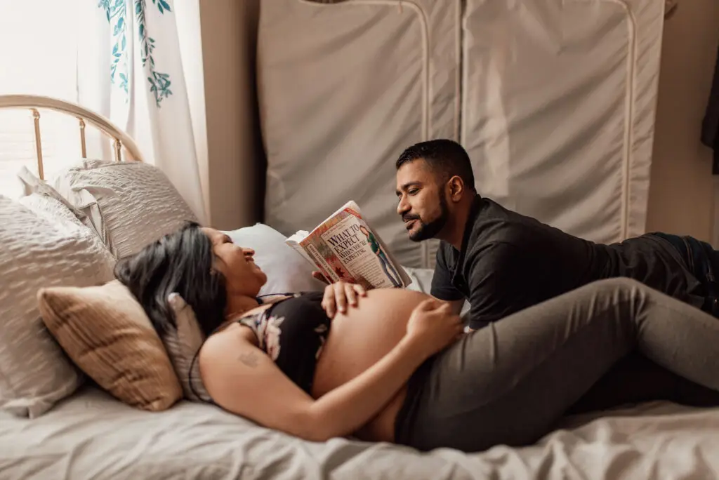 Brown couple laughing together. Pregnant woman laying down, gazing up at male partner reading to her.