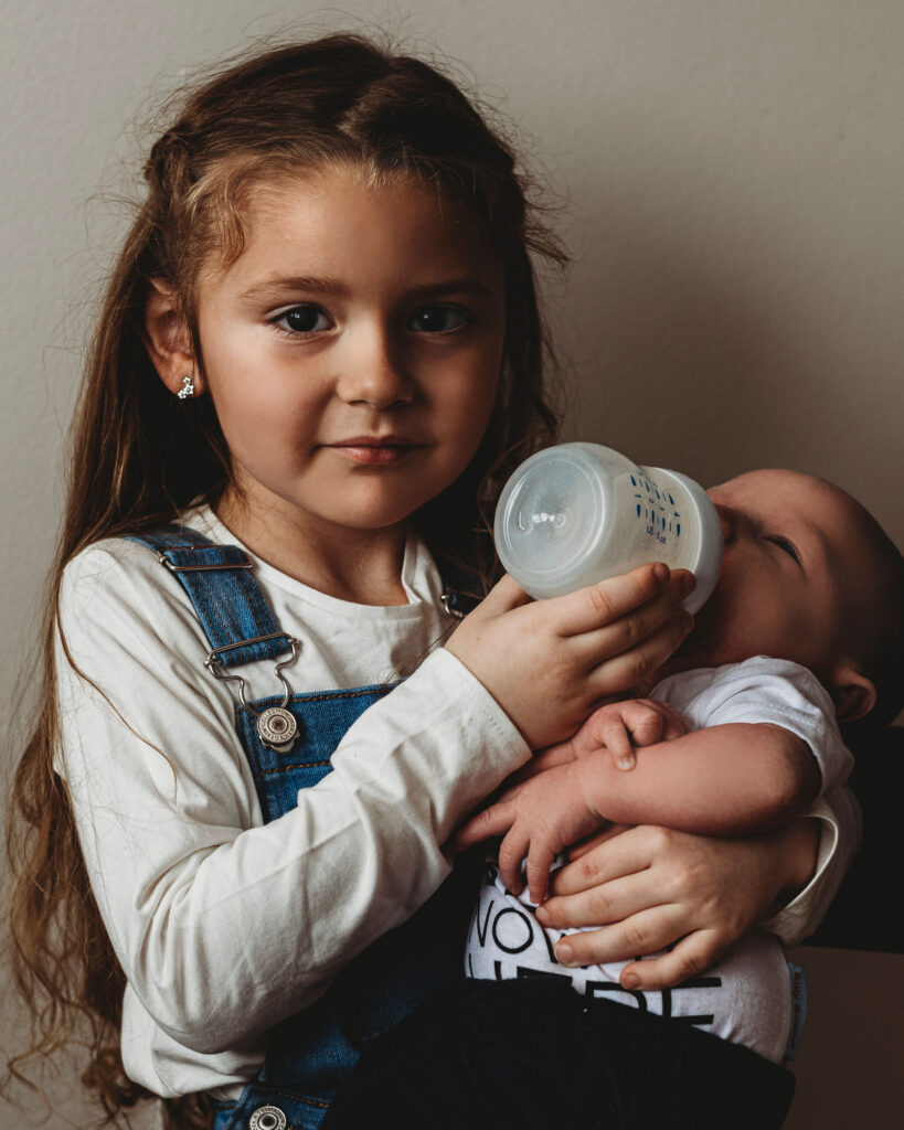 A young girl with dark hair stares at the camera holding a baby whom she is bottle feeding in an elevated position.